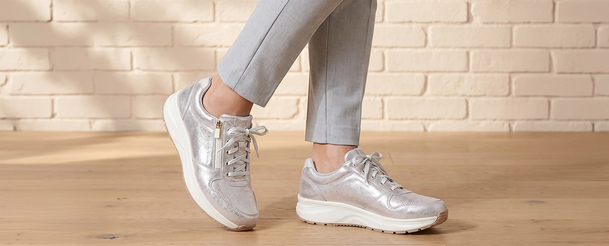 Model trägt silberne Damen-Sneaker mit Reißverschluss, Schnürung und weißer Komfortsohle auf hellem Holzboden