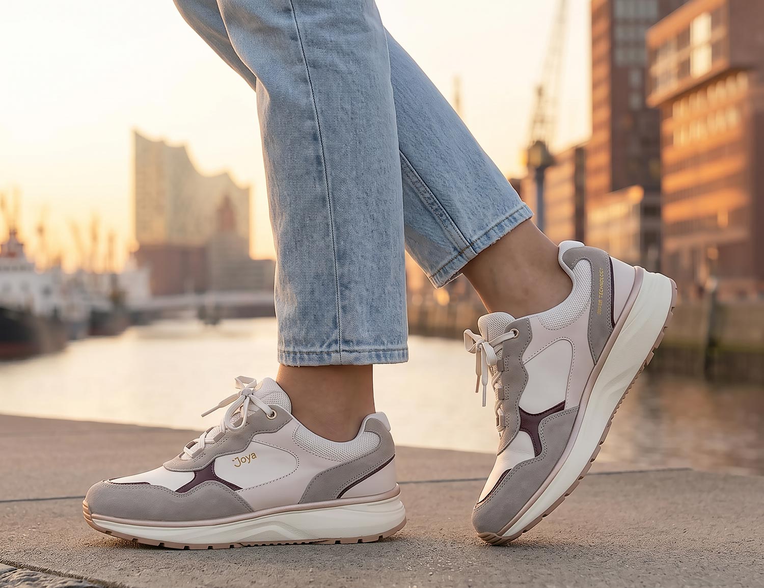 Frau trägt den JOYA Tina III beige-red, einen beigen Damen-Sneaker mit roten Akzenten und komfortabler breiter Passform, beim Spaziergang am Wasser in der Stadt.