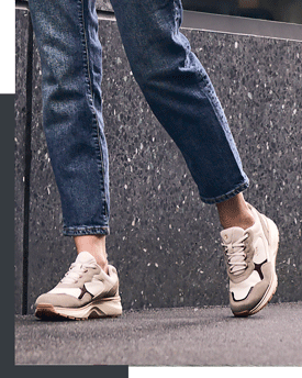 Person trägt weiße und beige Sneaker mit dicker Sohle und blauer Jeans auf grauem Boden vor dunkler Steinwand