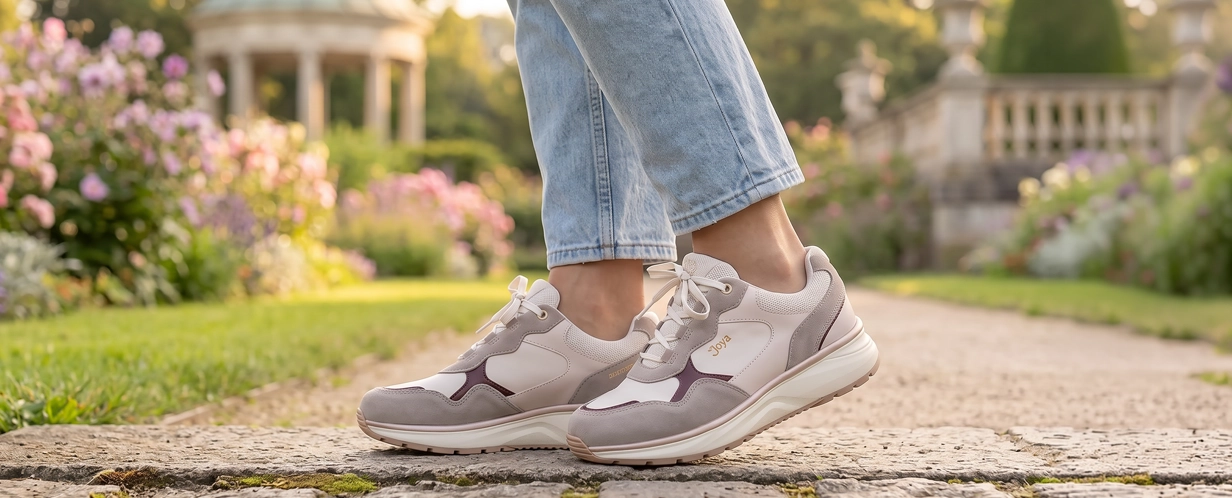 Damen tragen weiße und beige Sneaker mit komfortabler Laufsohle auf einem steinigen Gartenweg vor blühender Parkkulisse