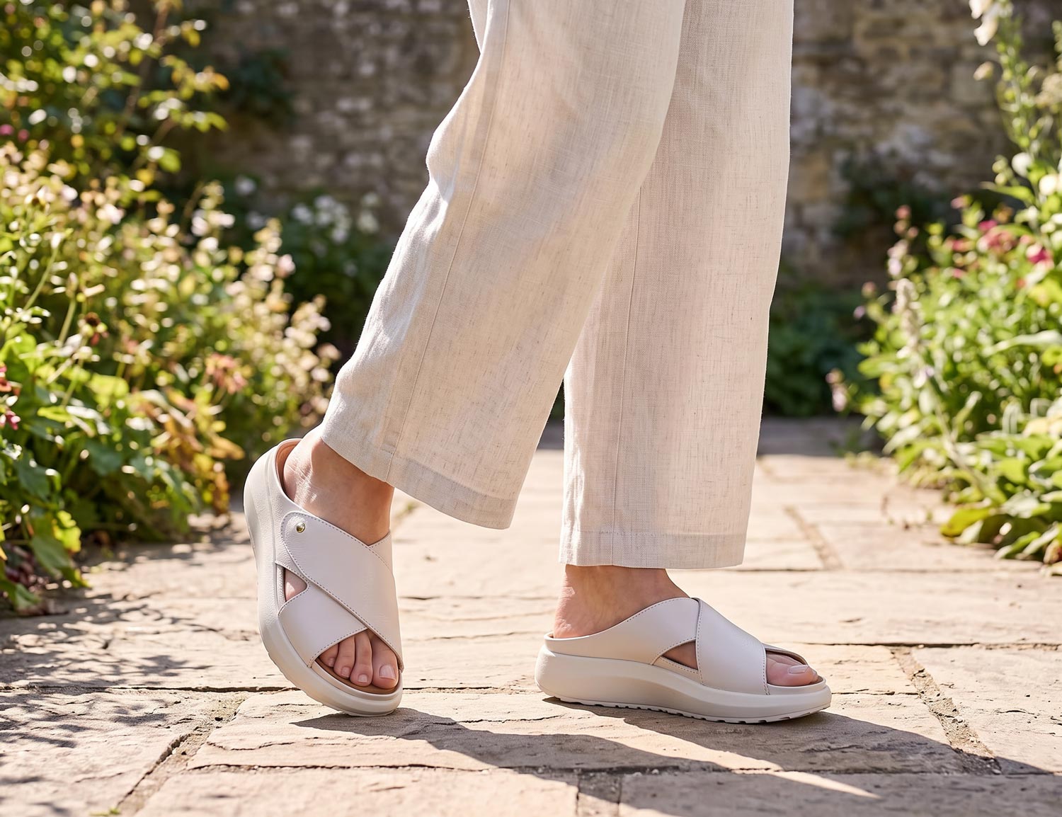 Dame trägt die JOYA Alicante W light beige, eine hellbeige Damen-Pantolette mit gekreuzten Riemen und bequemer Keilsohle, auf einem Gartenweg.