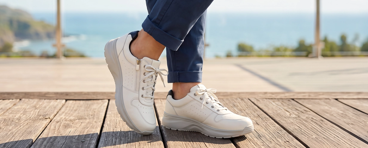 Nahaufnahme weißer Damen-Sneaker mit Reißverschluss, Schnürung und sportlicher Sohle, getragen auf einer Holzterrasse mit Meerblick